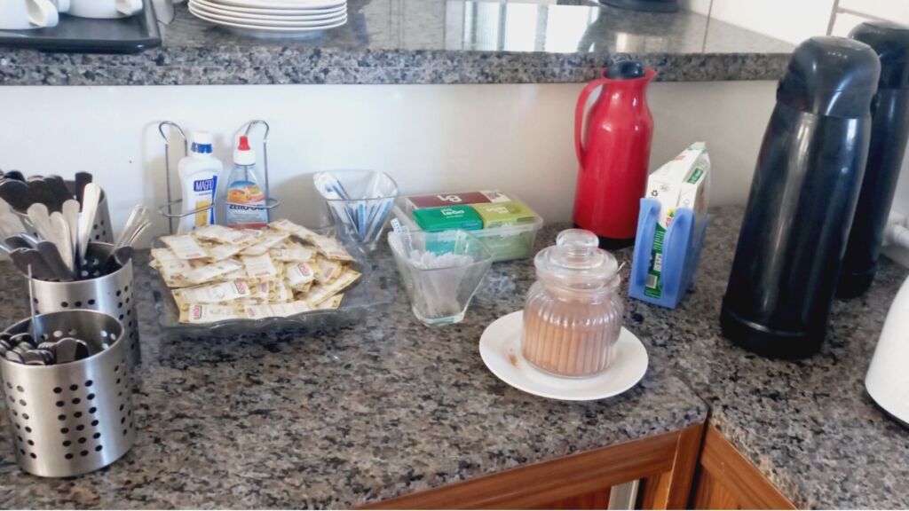 Balcão de café com garrafas térmicas, sachês de adoçante e utensílios organizados.