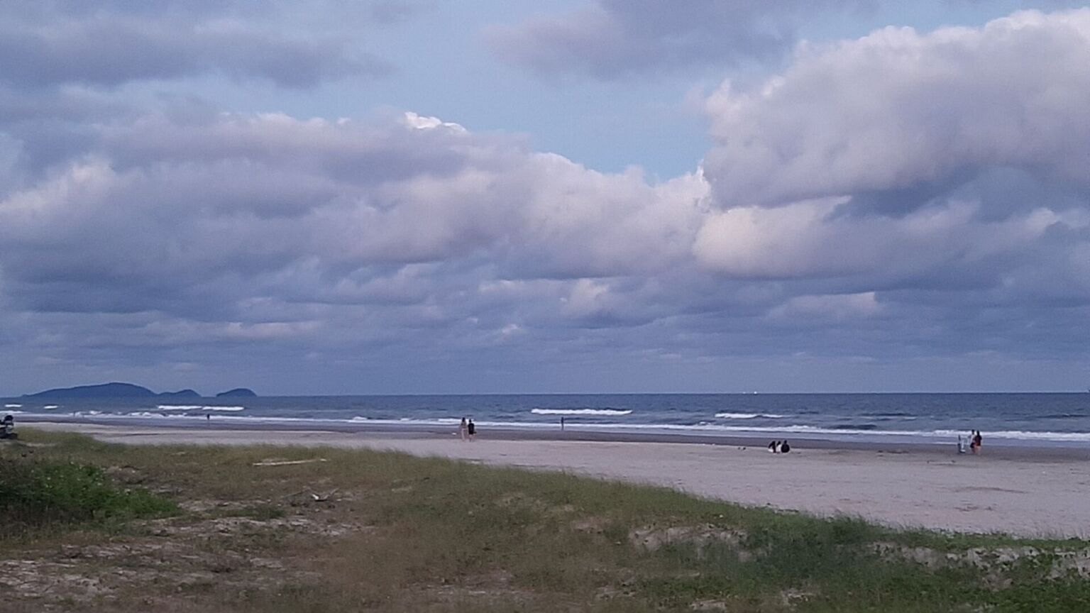 Praia com areia escura, céu nublado e ondas suaves ao fundo, poucas pessoas caminhando.