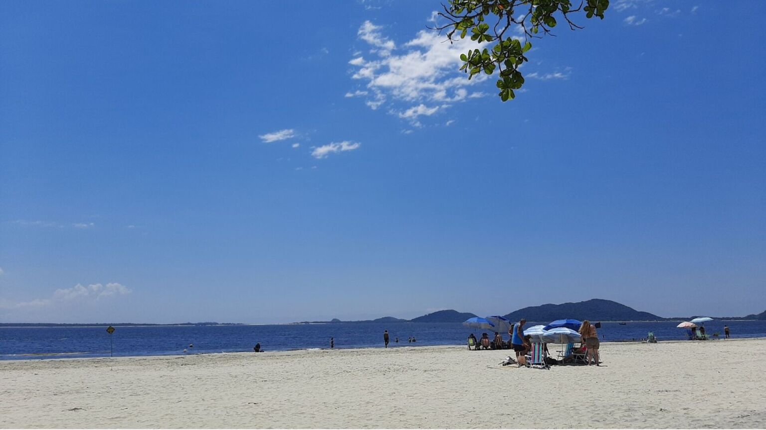 Praia de areia clara com céu azul e poucas nuvens, pessoas sentadas sob guarda-sóis próximos ao mar calmo.